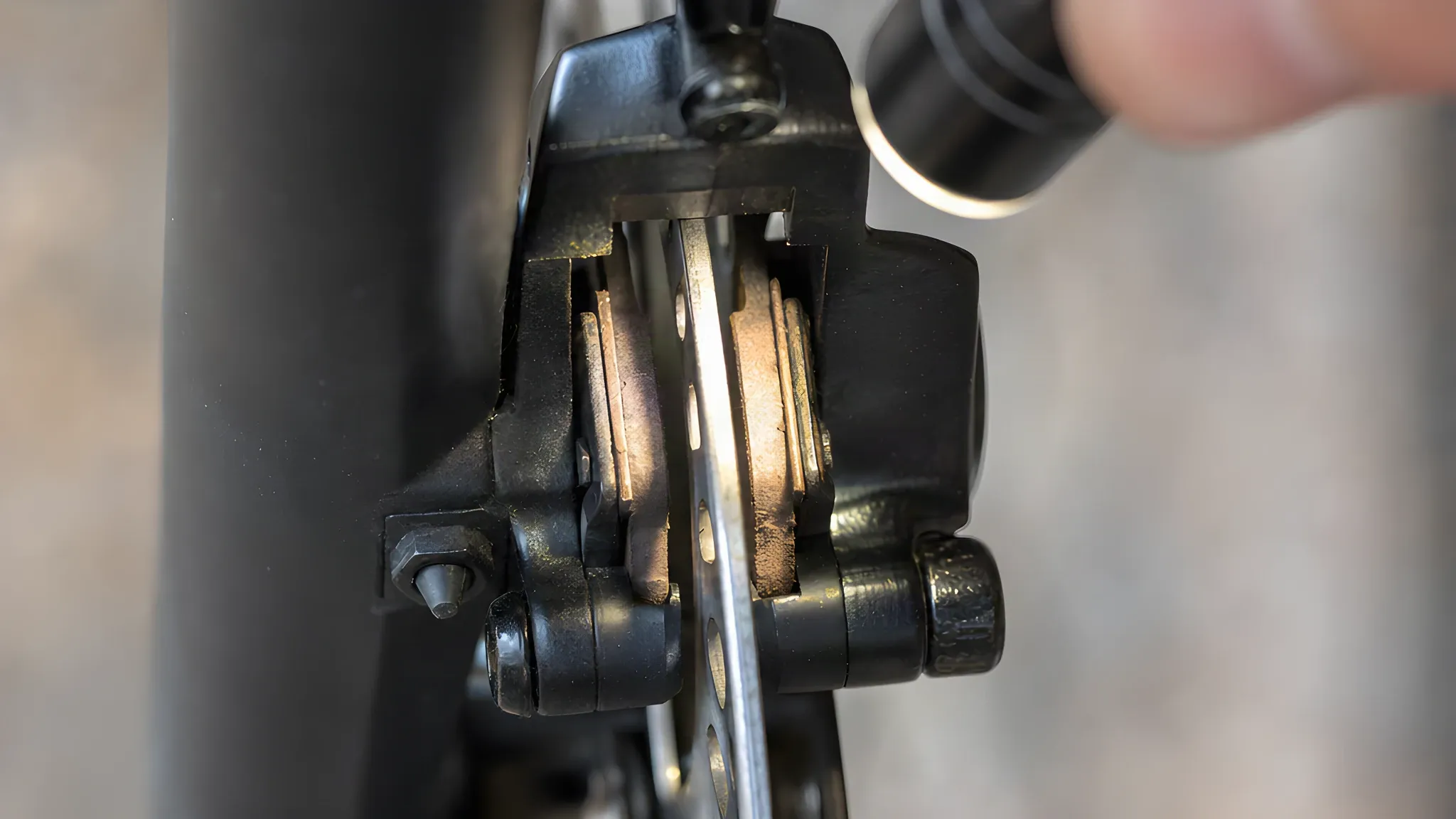 Close-up of a hydraulic disc brake calliper lit by a small torch, the rotor running between two brake pads with their pad material visible