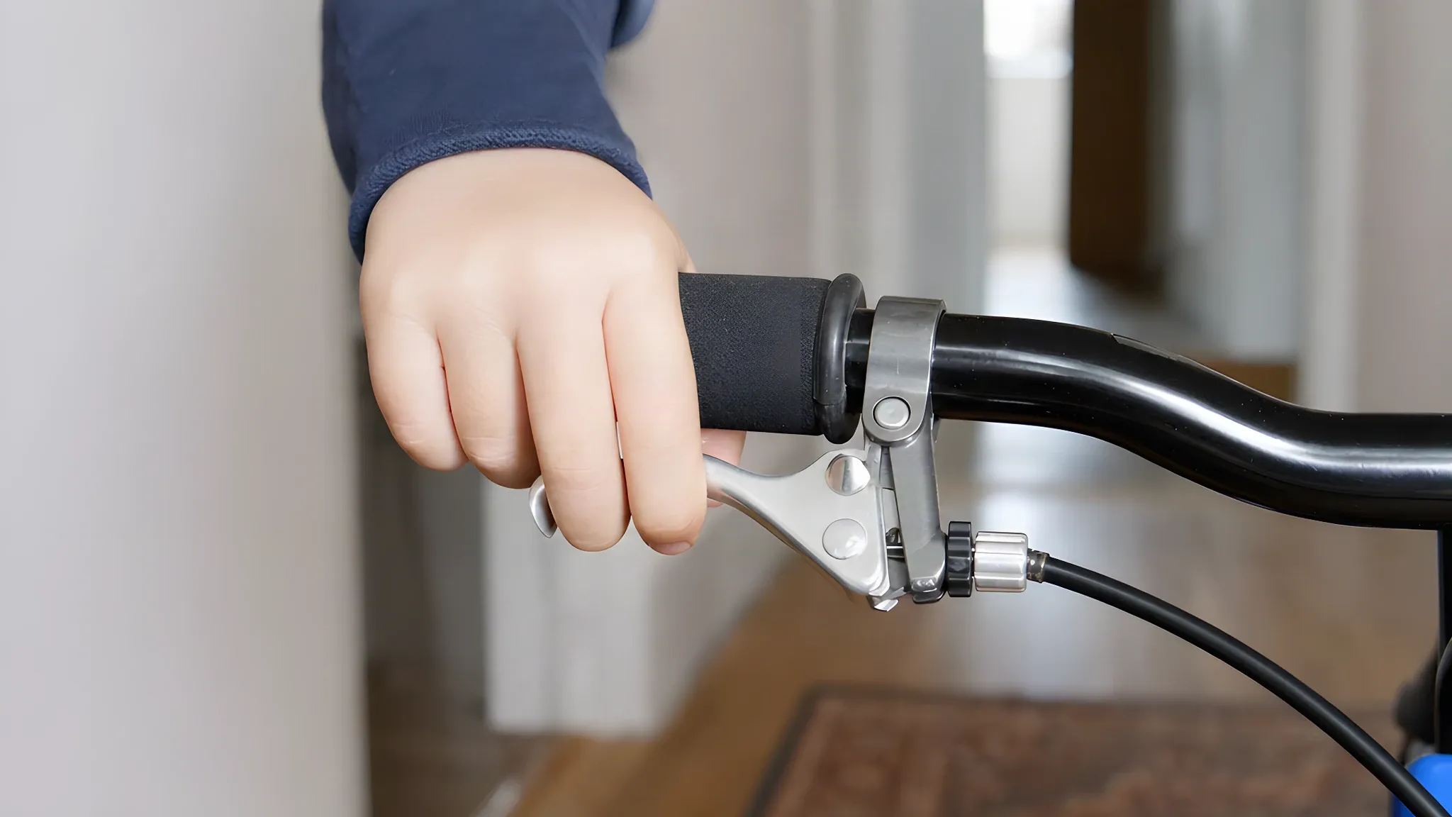 A child's hand on the foam grip of a small bike's flat handlebar, fingers on a short alloy brake lever with the cable running from the lever body