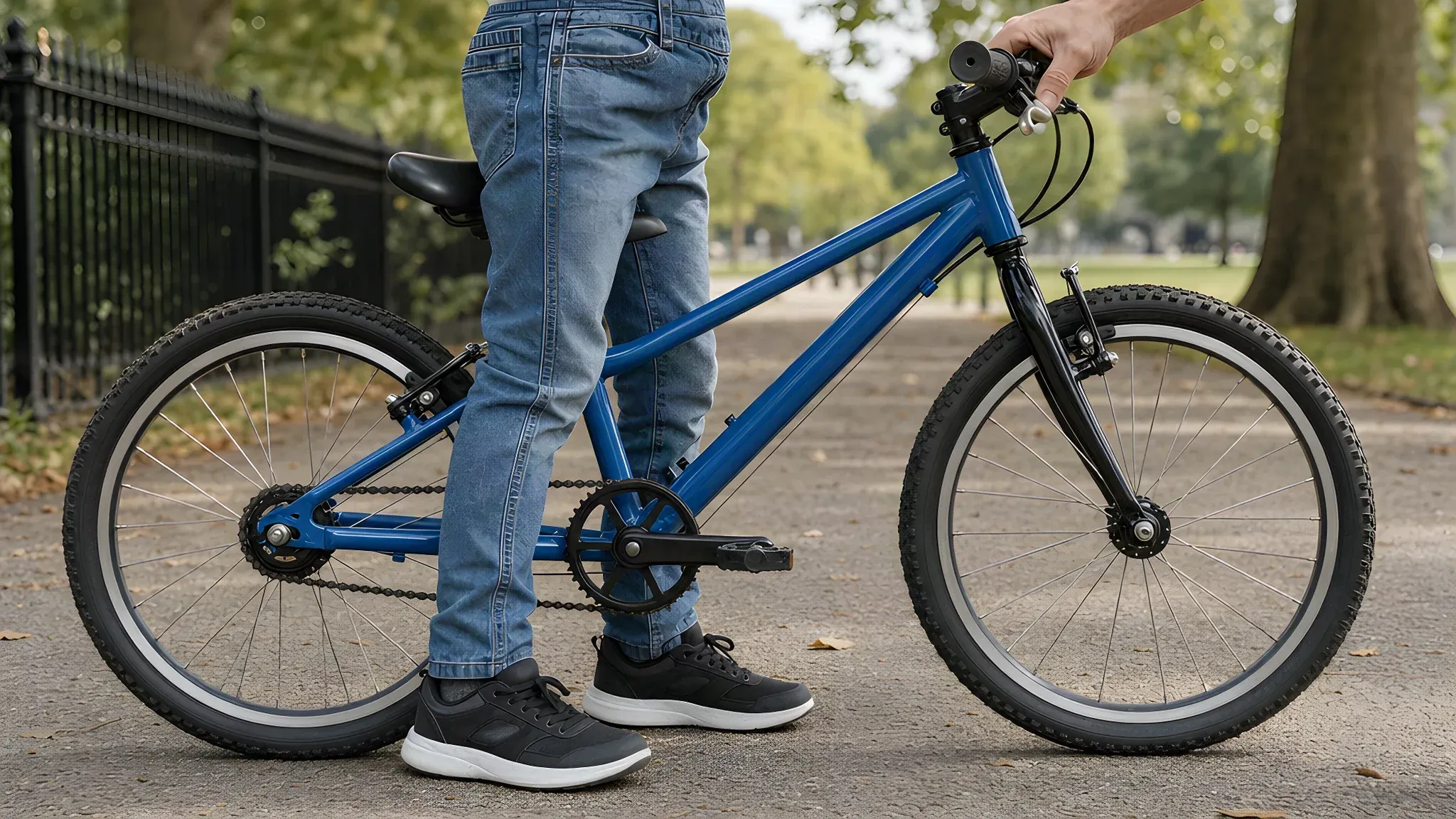 Child in jeans standing astride the top tube of a blue 20 inch bike on a park path with both feet flat, an adult hand steadying the handlebar