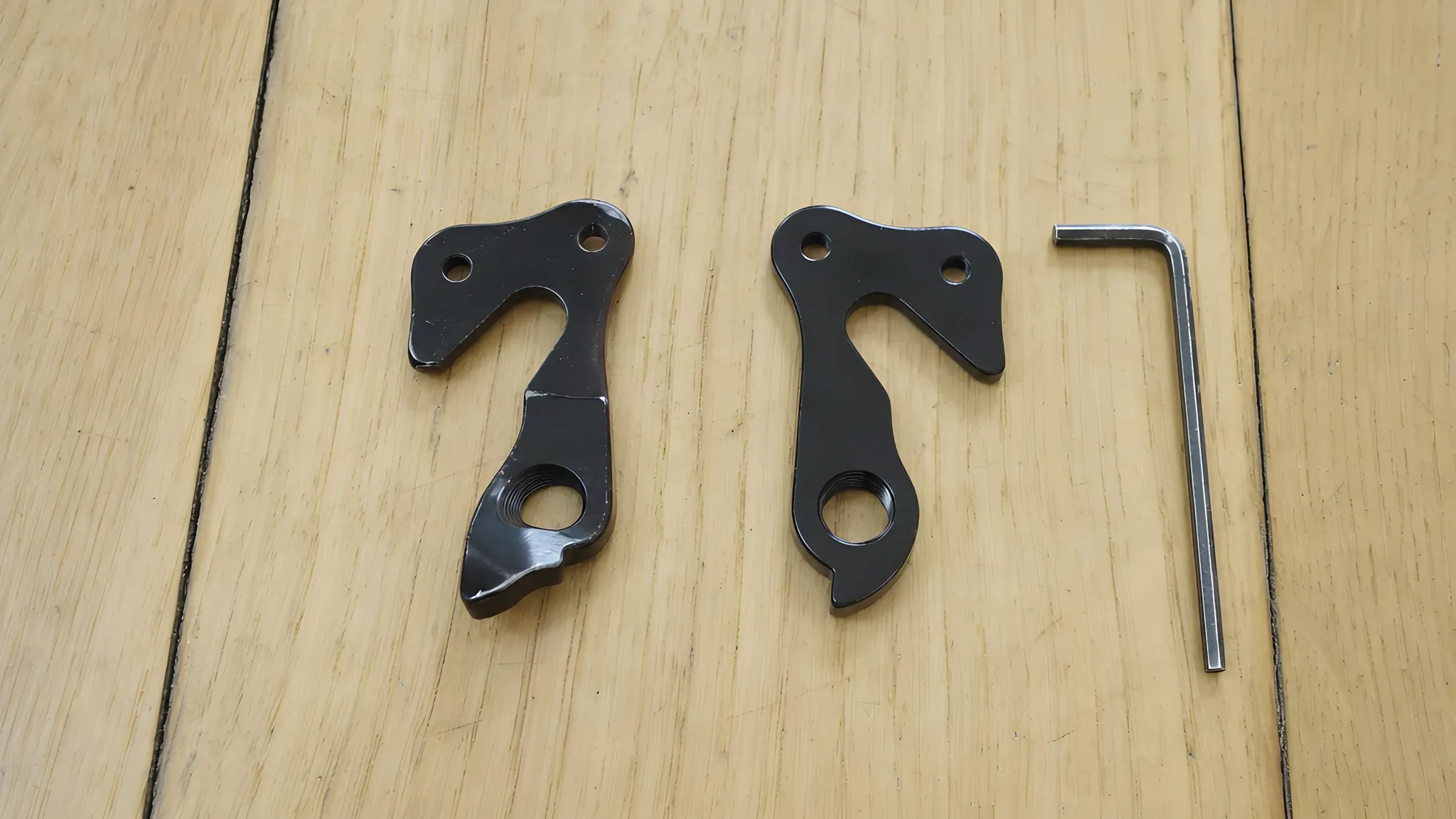 A bent black derailleur hanger next to a straight new hanger of the same shape and an Allen key, on a wooden hallway floor