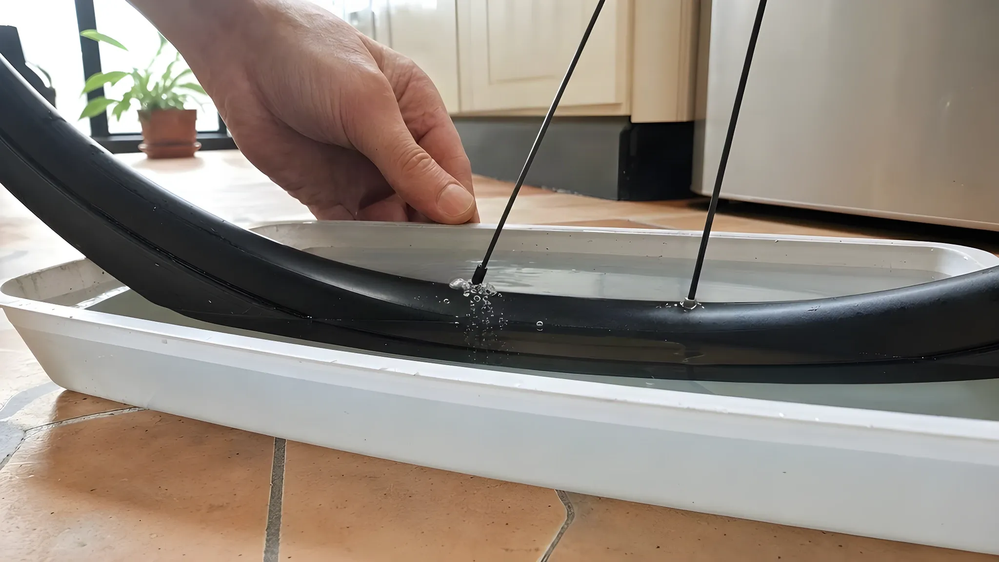 Lower part of a road bike wheel standing in a shallow tray of water on a kitchen floor, with small air bubbles rising from the submerged rim beside a spoke nipple