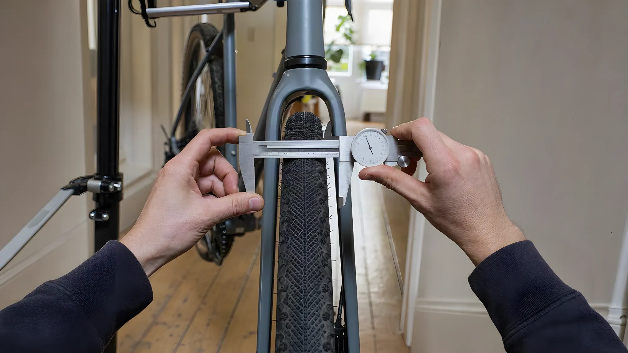 Two hands holding a dial calliper across the widest point of an inflated gravel bike tyre, with the front wheel still in the fork and the bike in a repair stand in a London hallway